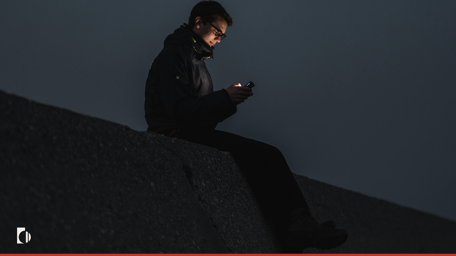 A figure sitting on a concrete ledge at night, lit only by their phone screen.